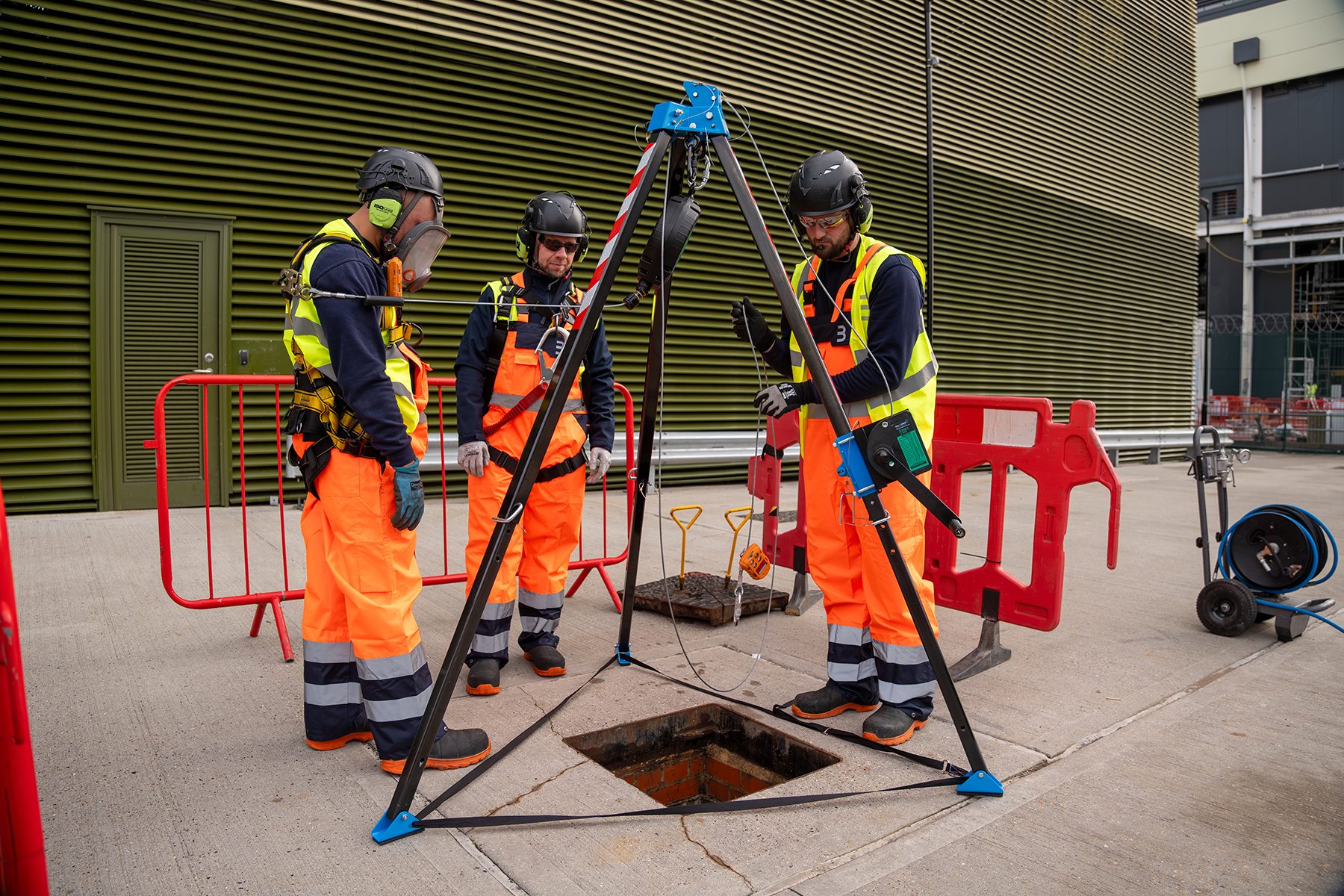 Three workers in safety gear and orange vests stand around an open manhole with tripod equipment during street maintenance