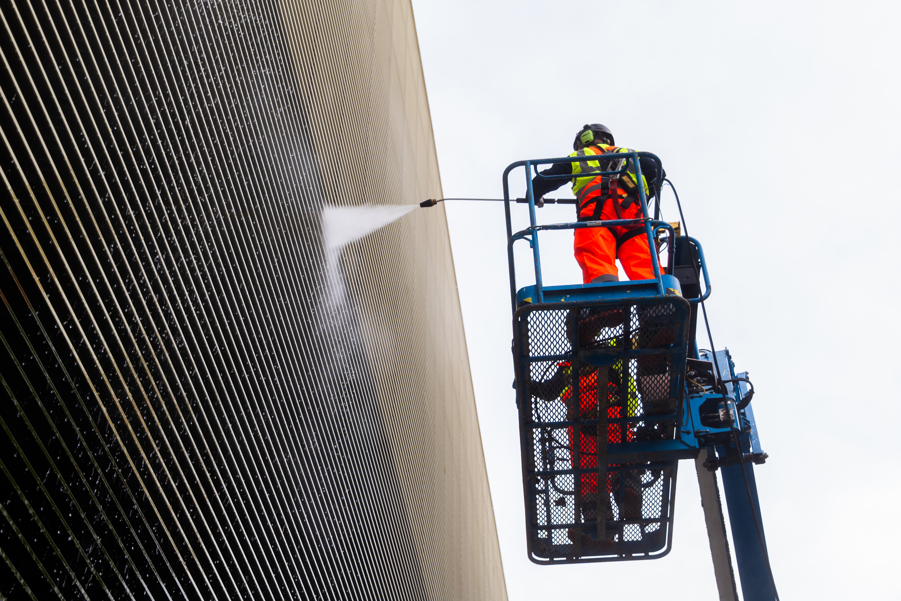 Worker in high-visibility gear spraying cleaning solution on corrugated metal building from elevated lift platform.