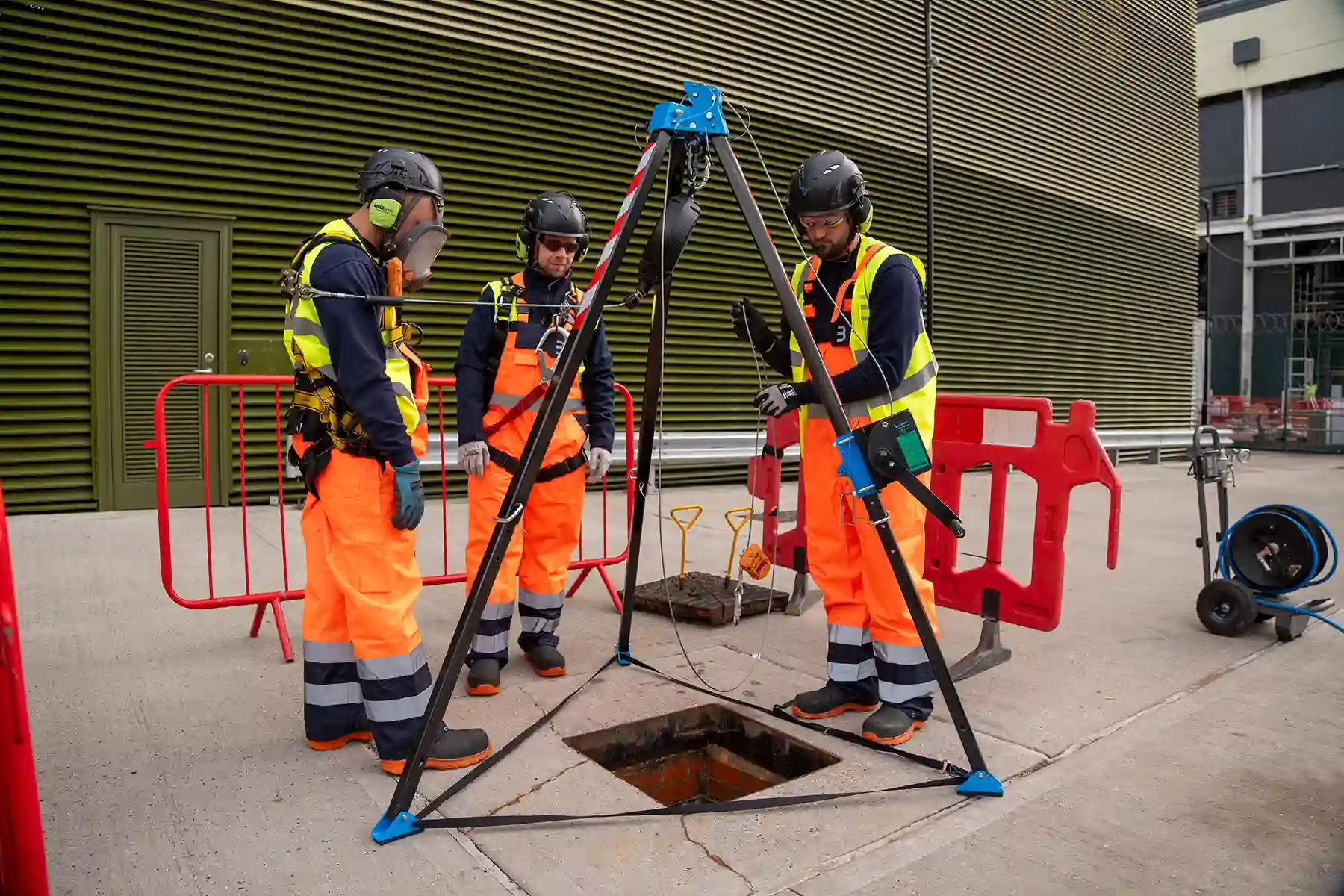 Three workers in safety gear and orange vests stand around an open manhole with tripod equipment during street maintenance