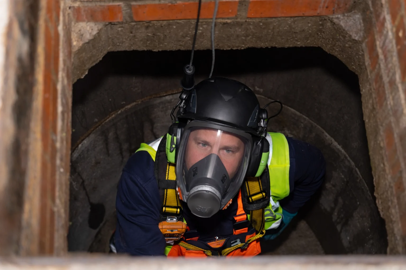 Worker in protective gear and respirator inside underground brick tunnel structure.
