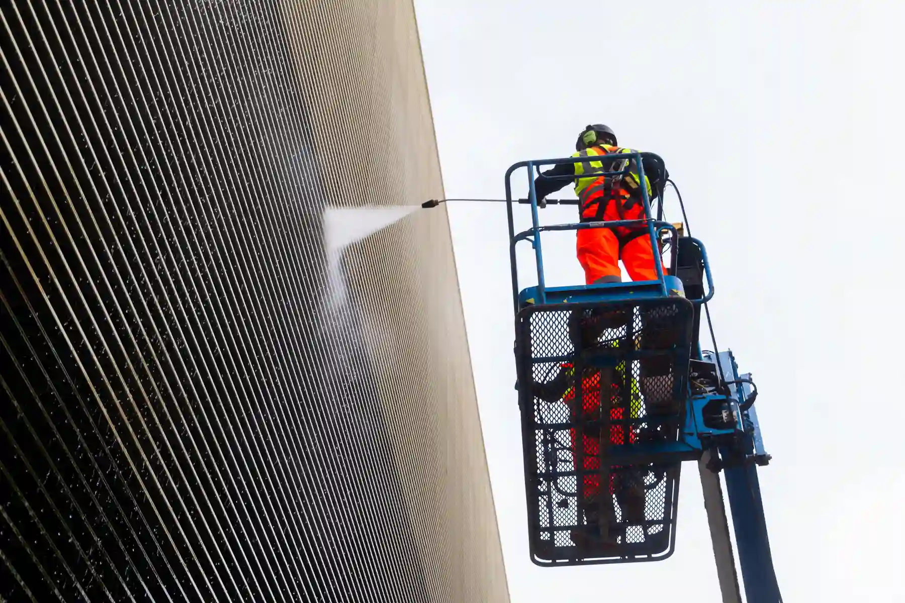 Worker in high-visibility gear spraying cleaning solution on corrugated metal building from elevated lift platform.