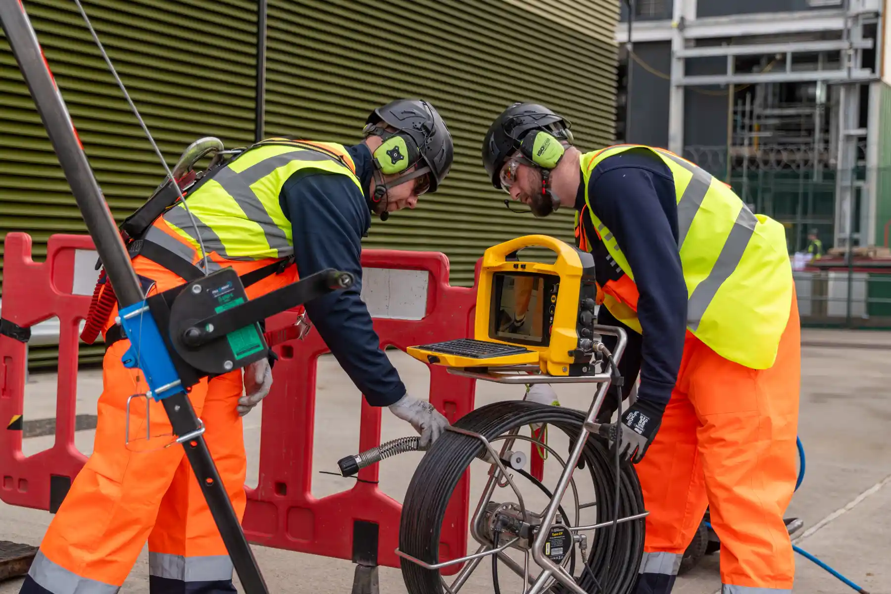 Two construction workers in high-visibility vests and helmets inspect underground infrastructure using diagnostic equipment