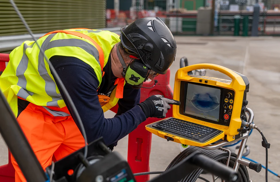 Worker in high-visibility vest and black helmet operating yellow survey equipment on industrial site.