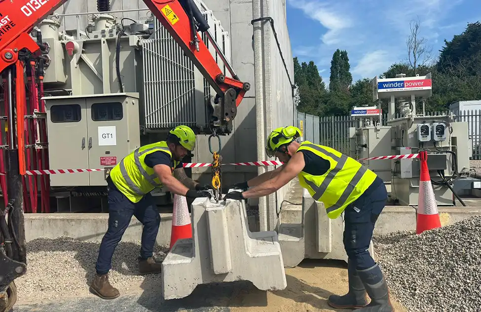 Two construction workers in yellow safety vests guiding a concrete barrier being lifted by a red crane at a work site.