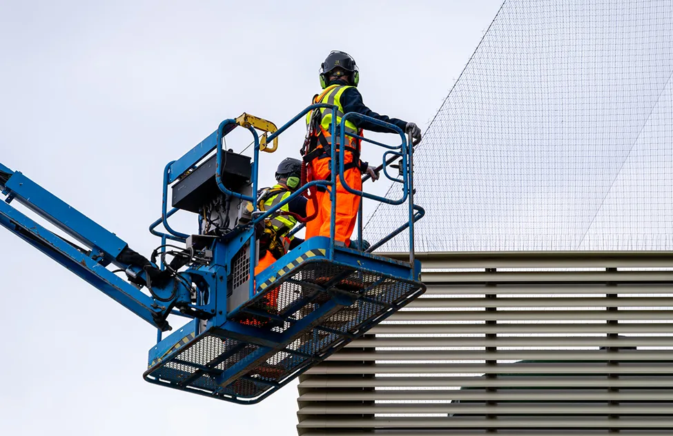 Two construction workers in safety gear operating a blue aerial lift platform against a modern building facade.