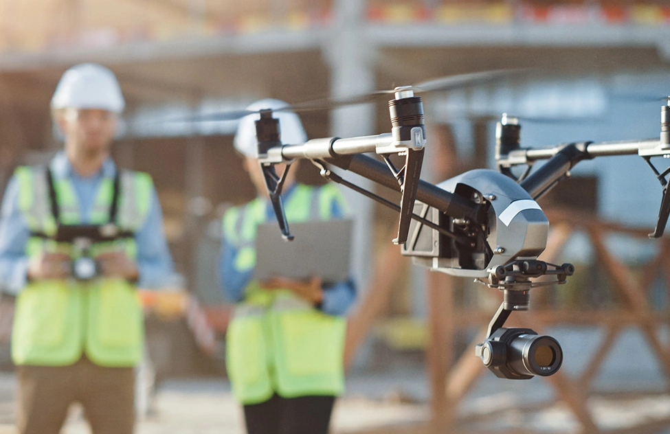 A drone hovers above a construction site while workers in safety vests and hard hats operate it.
