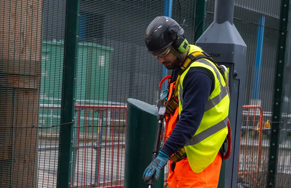 Worker in yellow safety vest and black helmet uses power drill on metal fence structure.